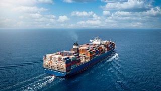 Großes Containerschiff auf offener See bei Tageslicht, beladen mit zahlreichen, bunt gestapelten Frachtcontainern. Das Schiff fährt von der Kamera weg in Richtung Horizont, hinterlässt eine weiße Bugwelle im tiefblauen Wasser. Der Himmel ist leicht bewölkt.