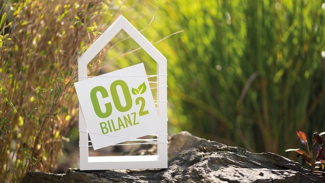 Symbolische Darstellung einer CO₂-Bilanz: Ein Schild in Form eines Hauses mit der Aufschrift ‚CO₂ Bilanz‘ steht auf einem Stein im Grünen.