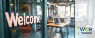 Glaswand mit dem Wort ‚Welcome‘ in großen Buchstaben; daneben das Logo des Welcome Center KMU Rheinisches Revier mit den Buchstaben ‚WCR‘ und dem Text ‚Welcome Center KMU Rheinisches Revier‘ auf unscharfem Bürohintergrund.