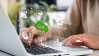 Das Bild zeigt eine Person, die auf einem Laptop tippt. Über der Tastatur sind transparente, schwebende Symbole zu sehen, die Nachhaltigkeit und Umweltschutz symbolisieren. Dazu gehören ein grünes CO₂-Reduktionssymbol, ein Recycling-Symbol, Solarenergie, Windkraft sowie ein Icon für nachhaltige Produktion. Die Szene wirkt futuristisch und digital, mit einer warmen Lichtstimmung im Hintergrund.