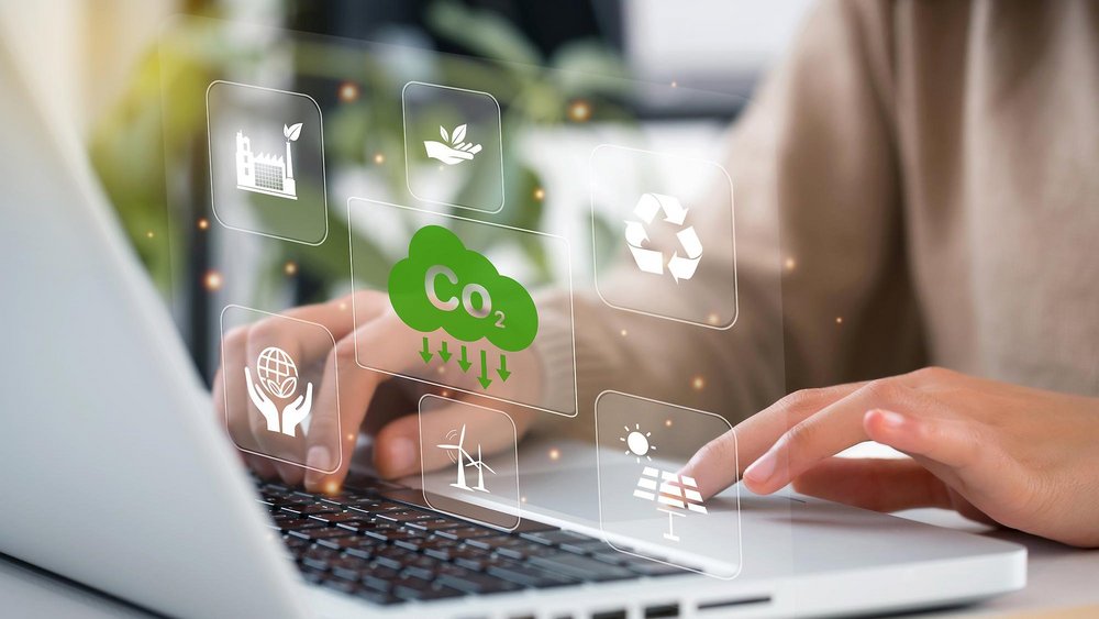 Das Bild zeigt eine Person, die auf einem Laptop tippt. Über der Tastatur sind transparente, schwebende Symbole zu sehen, die Nachhaltigkeit und Umweltschutz symbolisieren. Dazu gehören ein grünes CO₂-Reduktionssymbol, ein Recycling-Symbol, Solarenergie, Windkraft sowie ein Icon für nachhaltige Produktion. Die Szene wirkt futuristisch und digital, mit einer warmen Lichtstimmung im Hintergrund.