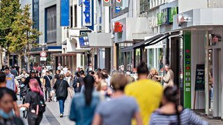 Belebte Einkaufsstraße in der Krefelder Innenstadt mit vielen Passanten. Ältere Menschen im Vordergrund, jüngere im Hintergrund. Läden mit auffälligen Werbeschildern säumen die Straße. Rechts im Bild ein Café mit roten Sonnenschirmen. Die Szene zeigt geschäftiges, urbanes Alltagsleben.