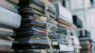 Stapel von dicht gefüllten Aktenordnern und Dokumentenmappen, die mit Bändern zusammengebunden sind, in einem Büro oder Archiv.