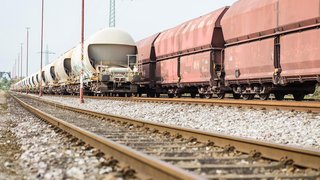 Ein langer Güterzug steht auf einem Bahngleis. Die Waggons haben unterschiedliche Formen und Farben – einige sind rostrot und eckig, andere sind hell und haben eine abgerundete, kuppelförmige Struktur. Der Zug erstreckt sich in die Ferne, während entlang der Gleise rote Beleuchtungsmasten stehen. Im Hintergrund sind Stromleitungen und eine blasse Himmel zu sehen. Der Boden besteht aus Schotter und Metallschienen.