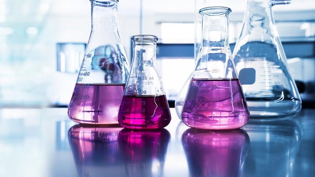 Das Bild zeigt mehrere Erlenmeyerkolben auf einer reflektierenden Laborarbeitsfläche. Einige der Kolben enthalten eine auffällig pink-violette Flüssigkeit, während andere mit einer klaren Substanz gefüllt sind. Der Hintergrund ist hell und verschwommen, was eine moderne Laborumgebung andeutet. Die Lichtreflexionen auf den Gefäßen und der Oberfläche erzeugen eine futuristische und sterile Atmosphäre.