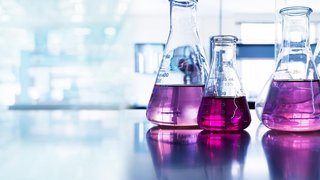 Das Bild zeigt mehrere Erlenmeyerkolben auf einer reflektierenden Laborarbeitsfläche. Einige der Kolben enthalten eine auffällig pink-violette Flüssigkeit, während andere mit einer klaren Substanz gefüllt sind. Der Hintergrund ist hell und verschwommen, was eine moderne Laborumgebung andeutet. Die Lichtreflexionen auf den Gefäßen und der Oberfläche erzeugen eine futuristische und sterile Atmosphäre.