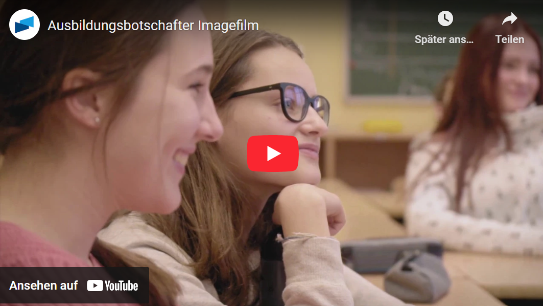 Zwei junge Frauen sitzen nebeneinander in einem Klassenzimmer und lächeln, während sie aufmerksam nach vorne blicken. Im Hintergrund sind weitere Schüler:innen sowie eine grüne Tafel zu sehen. Das Bild stammt aus einem YouTube-Video mit dem Titel „Ausbildungsbotschafter Imagefilm“, das in der oberen linken Ecke eingeblendet ist. In der Mitte befindet sich ein roter Play-Button von YouTube.
