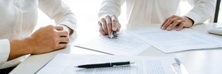 Zwei Personen in weißer Kleidung sitzen an einem Schreibtisch und besprechen gemeinsam Dokumente mit Tabellen und Diagrammen; eine Person zeigt mit einem Stift auf ein Balkendiagramm.