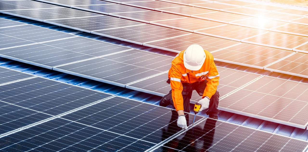 Ein Techniker in orangefarbener Schutzkleidung und Helm installiert oder wartet Solarmodule auf einem Dach bei Sonnenschein.