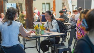 Azubi-Speed-Dating im Gare du Neuss: Eine junge Frau sitzt lächelnd mit Unterlagen an einem Tisch in einem belebten Innenraum und führt ein Gespräch mit einer anderen Person; um sie herum sitzen weitere Menschen an Tischen.