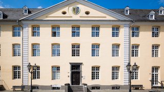 Das Bild zeigt das Rathaus von Brüggen, ein großes, hellgelb gestrichenes, dreistöckiges Gebäude mit vielen weißen Sprossenfenstern und einem dunklen Holzeingang in der Mitte. Über dem Eingang befindet sich ein Dreiecksgiebel mit dem Wappen der Gemeinde Brüggen. Vor dem Gebäude stehen zwei alte Straßenlaternen und Holzbänke auf einem gepflasterten Platz. Der Himmel ist klar und blau, was dem Bild eine helle und freundliche Stimmung verleiht.