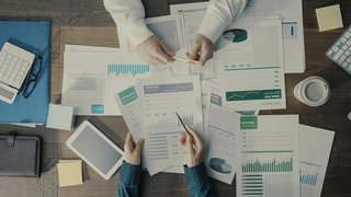 Schreibtischszene aus der Vogelperspektive: Zwei Personen analysieren gemeinsam Finanzdokumente mit Diagrammen und Tabellen. Auf dem Tisch liegen ein Taschenrechner, eine Brille, ein Tablet, ein Kaffeebecher, ein Notizbuch, Klebezettel und eine Tastatur. Die Szene wirkt wie eine konzentrierte Büroarbeit oder Besprechung.