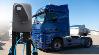 Elektroladesäule im Vordergrund mit Ladekabeln; im Hintergrund steht ein blauer Lkw auf einem Betriebshof.