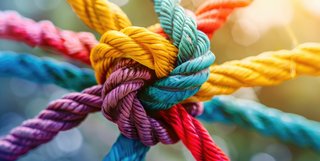 Nahaufnahme eines Knotens aus mehreren bunten Seilen, die in verschiedenen Farben wie Gelb, Rot, Blau, Grün, Lila und Orange fest miteinander verflochten sind; symbolisiert Stärke, Zusammenarbeit und Vielfalt.