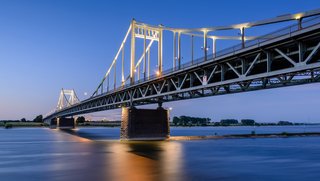 Beleuchtete Uerdinger Rheinbrücke bei Dämmerung über dem Rhein, mit reflektierendem Wasser und blauem Himmel im Hintergrund. Die Brücke ist aus Stahl und zeigt eine markante Tragstruktur mit mehreren Pfeilern im Wasser.