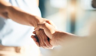 Zwei Personen reichen sich die Hände zu einem respektvollen Handschlag. Der Fokus liegt auf den Händen im Vordergrund, während die Personen im Hintergrund unscharf bleiben. Das Bild vermittelt Vertrauen, Zusammenarbeit und zwischenmenschliche Verbindung.
