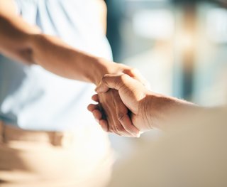Zwei Personen reichen sich die Hände zu einem respektvollen Handschlag. Der Fokus liegt auf den Händen im Vordergrund, während die Personen im Hintergrund unscharf bleiben. Das Bild vermittelt Vertrauen, Zusammenarbeit und zwischenmenschliche Verbindung.