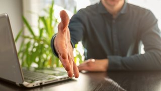 Ein Mann in dunklem Hemd sitzt an einem Schreibtisch mit Laptop und streckt freundlich die rechte Hand zur Begrüßung aus. Im Hintergrund sind eine grüne Pflanze und Tageslicht durch ein Fenster zu sehen. Die Szene vermittelt Offenheit und Gesprächsbereitschaft, beispielsweise in einem Bewerbungsgespräch oder beruflichen Meeting.