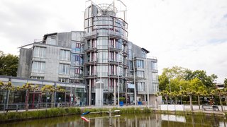 Das Bild zeigt das Rathaus von Kaarst. Das moderne Verwaltungsgebäude besteht aus mehreren Geschossen mit einer Fassade aus Glas- und Metallelementen. In der Mitte erhebt sich ein runder, verglaster Turm mit Balkonen. Vor dem Gebäude befindet sich ein Teich mit Schilf am Rand, in dem sich das Rathaus spiegelt. Bäume, Sitzgelegenheiten und ein gepflasterter Platz umgeben das Gebäude. Der Himmel ist hell und leicht bewölkt.