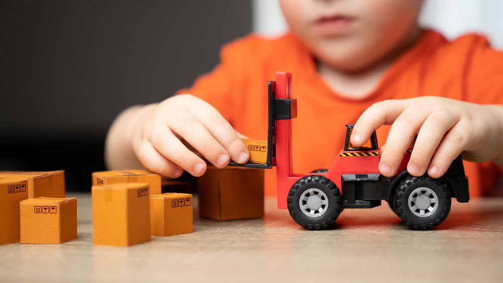Ein Kind spielt mit einem kleinen roten Spielzeug-Gabelstapler und hebt mehrere Miniatur-Pappkartons auf einem Tisch an.
