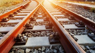 Nahaufnahme zweier paralleler Eisenbahnschienen, die auf einem Schotterbett verlegt sind und sich in der Ferne leicht auseinander bewegen. Die Szene ist in warmes Sonnenlicht getaucht, was den Schienen eine rötliche Färbung verleiht.