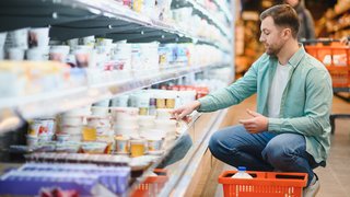 Ein Mann hockt in einem Supermarkt vor einem Kühlregal und nimmt einen Becher Joghurt aus dem unteren Fach, während ein Einkaufs­korb neben ihm steht.