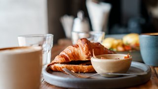 Nahaufnahme eines Frühstückstischs in einem Café. Auf einem Teller liegt ein frisch gebackenes, aufgeschnittenes Croissant neben einer kleinen Schale mit Aufstrich. Im Vordergrund steht eine Tasse mit Kaffee, im Hintergrund sind Gläser und weitere Teller leicht unscharf zu erkennen. Warmes Licht schafft eine gemütliche Atmosphäre.