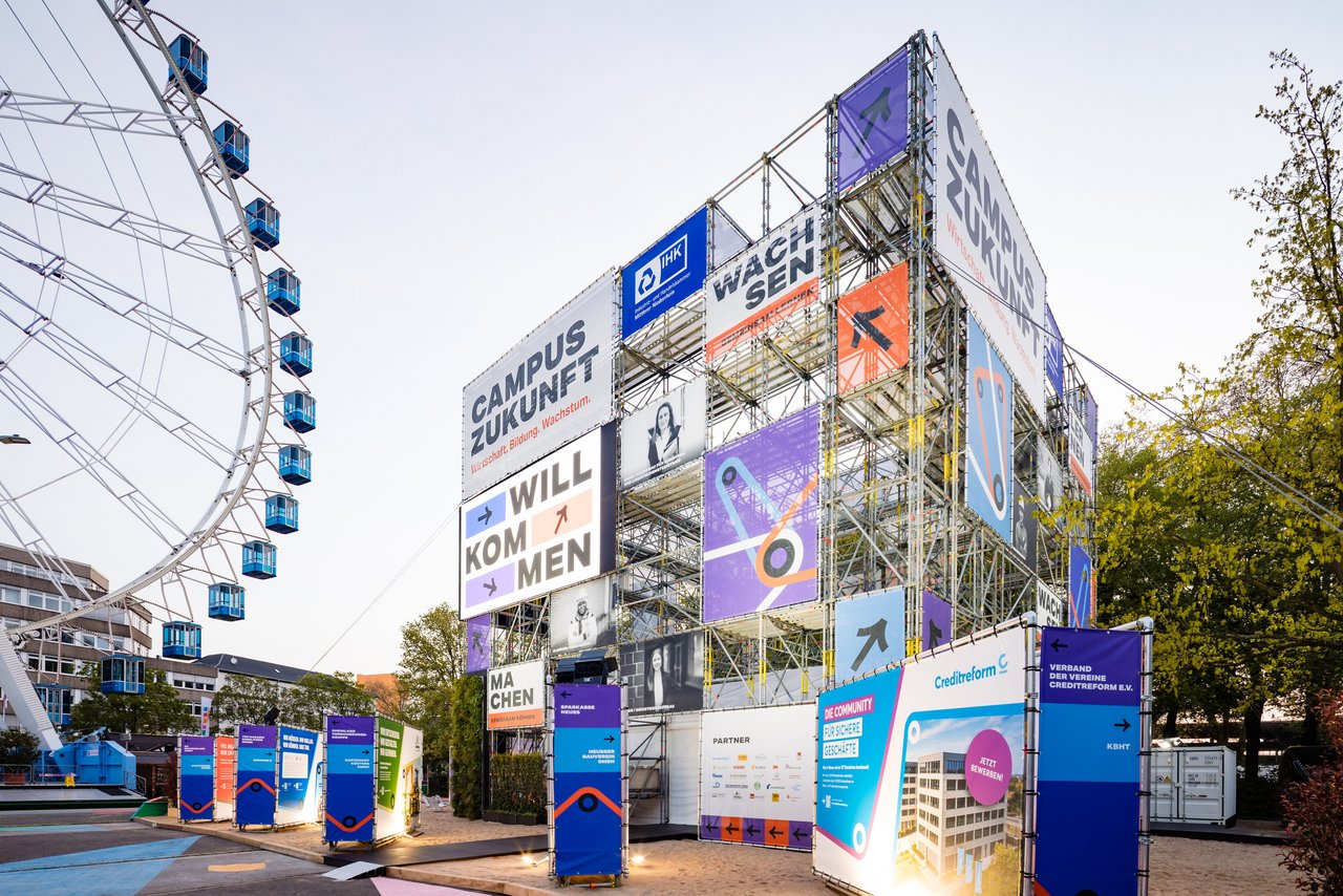 Außenansicht einer temporären Veranstaltungsarchitektur aus Gerüsten mit großen Beschilderungen wie „Campus Zukunft“, daneben ein Riesenrad auf einem öffentlichen Platz.