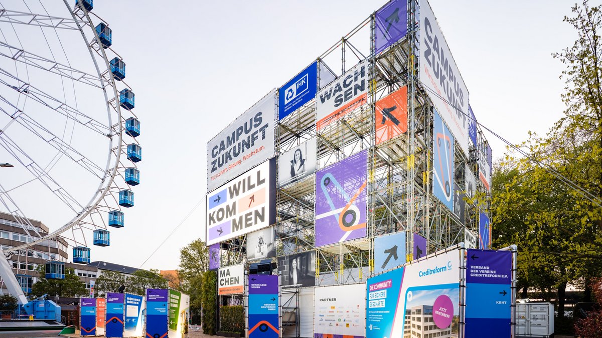 Außenansicht einer temporären Veranstaltungsarchitektur aus Gerüsten mit großen Beschilderungen wie „Campus Zukunft“, daneben ein Riesenrad auf einem öffentlichen Platz.