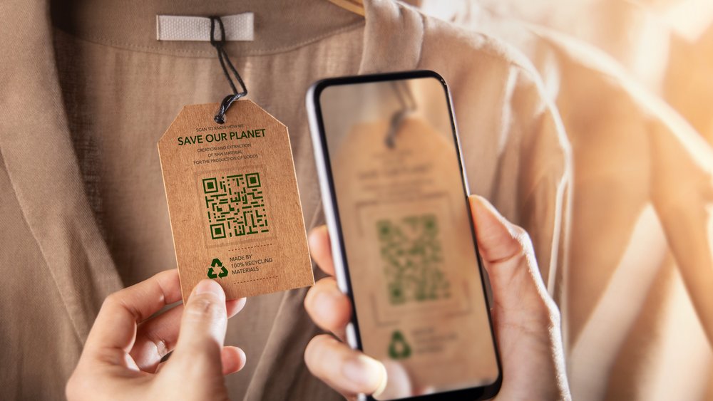 Eine Person hält ein nachhaltiges Bekleidungsetikett aus recyceltem Karton, das an einem beigen Kleidungsstück befestigt ist. Das Etikett enthält eine grüne QR-Code-Grafik und den Text "SAVE OUR PLANET" sowie Hinweise zur nachhaltigen Produktion. Die andere Hand hält ein Smartphone, das den QR-Code scannt. Die Szene ist in warmem Licht gehalten, was eine natürliche und umweltfreundliche Atmosphäre vermittelt.