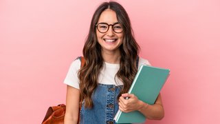 Lächelnde junge Frau mit langen braunen Haaren, Brille und Jeanskleid steht vor einem rosa Hintergrund. Sie trägt einen braunen Lederrucksack und hält Hefte oder Ordner in der Hand.