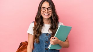 Lächelnde junge Frau mit langen braunen Haaren, Brille und Jeanskleid steht vor einem rosa Hintergrund. Sie trägt einen braunen Lederrucksack und hält Hefte oder Ordner in der Hand.