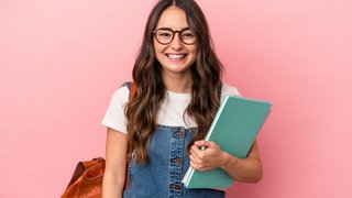 Lächelnde junge Frau mit langen braunen Haaren, Brille und Jeanskleid steht vor einem rosa Hintergrund. Sie trägt einen braunen Lederrucksack und hält Hefte oder Ordner in der Hand.