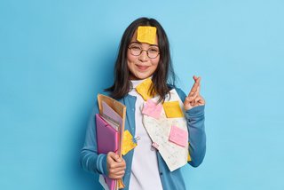 Junge Frau mit langen dunklen Haaren und runder Brille steht vor einem blauen Hintergrund. Sie trägt eine blaue Strickjacke und hält Ordner sowie Unterlagen im Arm. Mehrere Notizzettel und Blätter mit mathematischen Formeln sind an ihrer Kleidung und Stirn befestigt. Sie lächelt leicht und hält die Finger der rechten Hand zum Glück gekreuzt.
