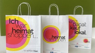 Drei weiße Papiertüten der Aktion „Heimat shoppen“ mit bunten Kreisdesigns und Aufschriften wie „Ich war heimat shoppen“ und „kauf lokal“.