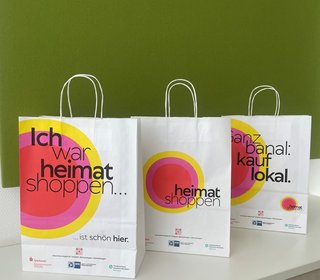 Drei weiße Papiertüten der Aktion „Heimat shoppen“ mit bunten Kreisdesigns und Aufschriften wie „Ich war heimat shoppen“ und „kauf lokal“.