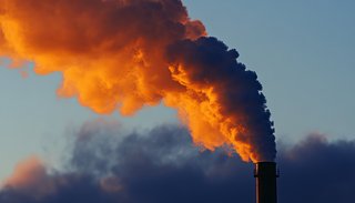 Ein Industrieschornstein stößt eine große Rauchwolke aus, die durch Sonnenlicht orangefarben leuchtet und sich kontrastreich vor dem blauen Himmel ausbreitet.