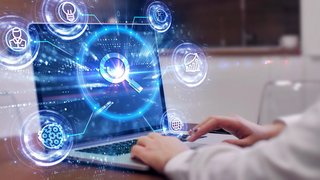 Person arbeitet an einem Laptop mit eingeblendeten digitalen Grafiken, darunter ein großes Lupensymbol für Suche und mehrere kreisförmige Icons für Ideen, Datenanalyse, Technologie und Vernetzung.