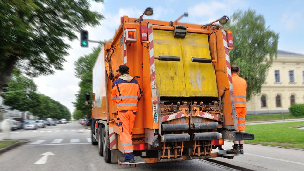 Zwei Müllwerker in orangefarbener Warnkleidung stehen auf Trittbrettern am Heck eines fahrenden Müllwagens. Der orangefarbene Müllwagen fährt bei grünem Ampellicht eine städtische Straße entlang. Bäume säumen die Straße auf der linken Seite, rechts ist ein helles, mehrstöckiges Gebäude mit Rundbogenfenstern zu sehen. Es ist Tag und der Himmel ist leicht bewölkt.