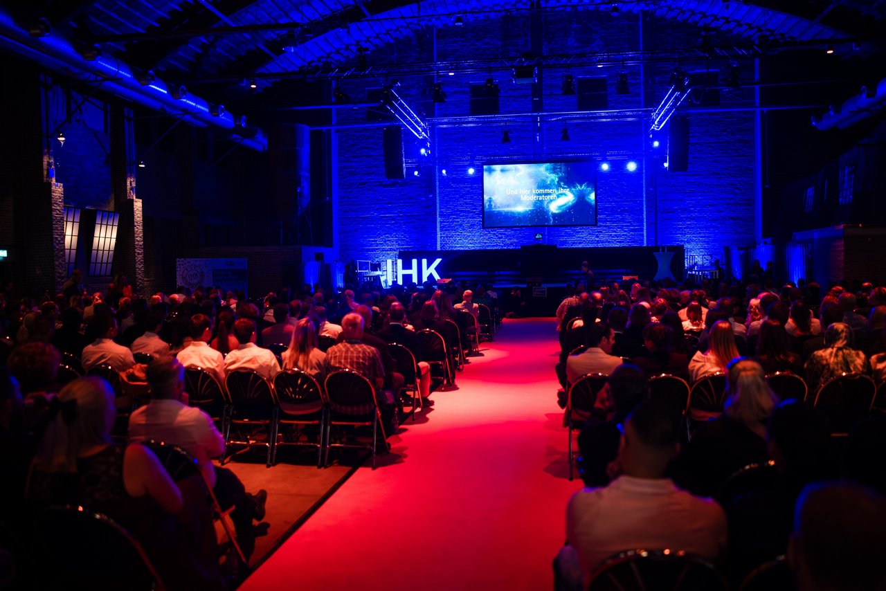 Bestenehrung 2025: : Publikum sitzt vor einer Bühne mit blauer Beleuchtung und schaut auf eine Präsentation mit dem IHK-Logo; ein roter Teppich führt durch den Mittelgang.