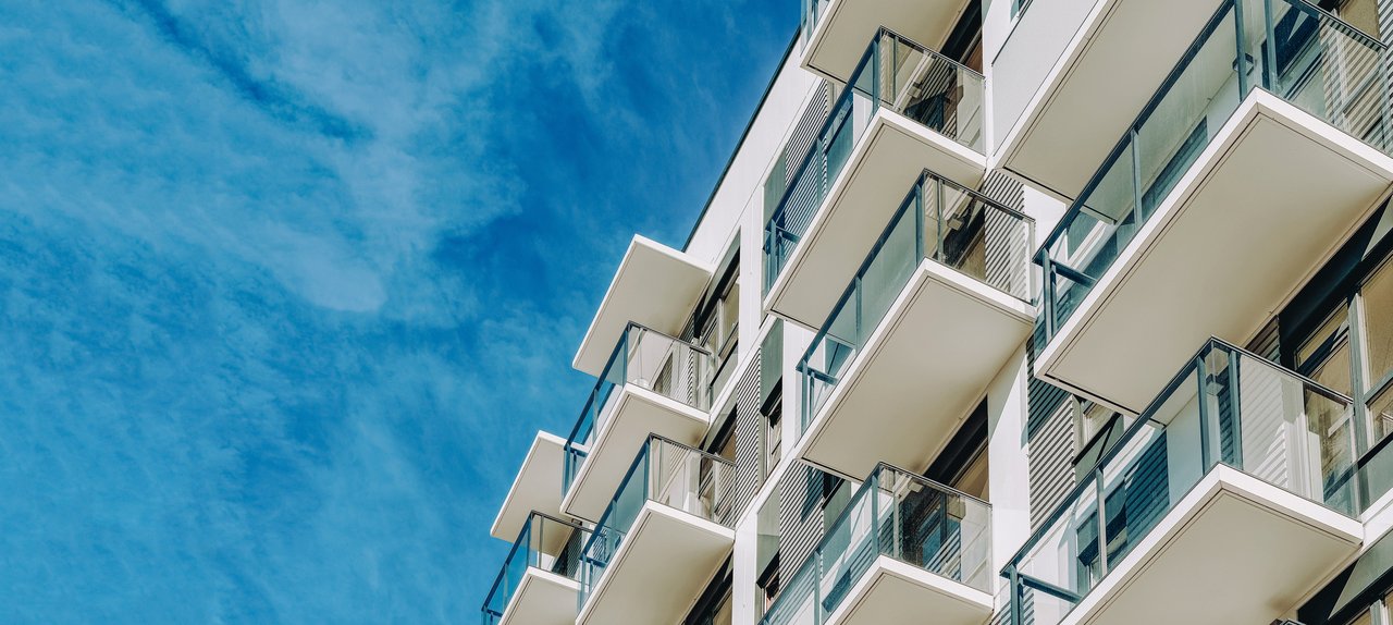 Ein modernes Wohngebäude mit mehreren Stockwerken und Balkonen, die in einem regelmäßigen Muster angeordnet sind. Die Balkone haben Glasgeländer und das Gebäude ist vor einem klaren blauen Himmel fotografiert.