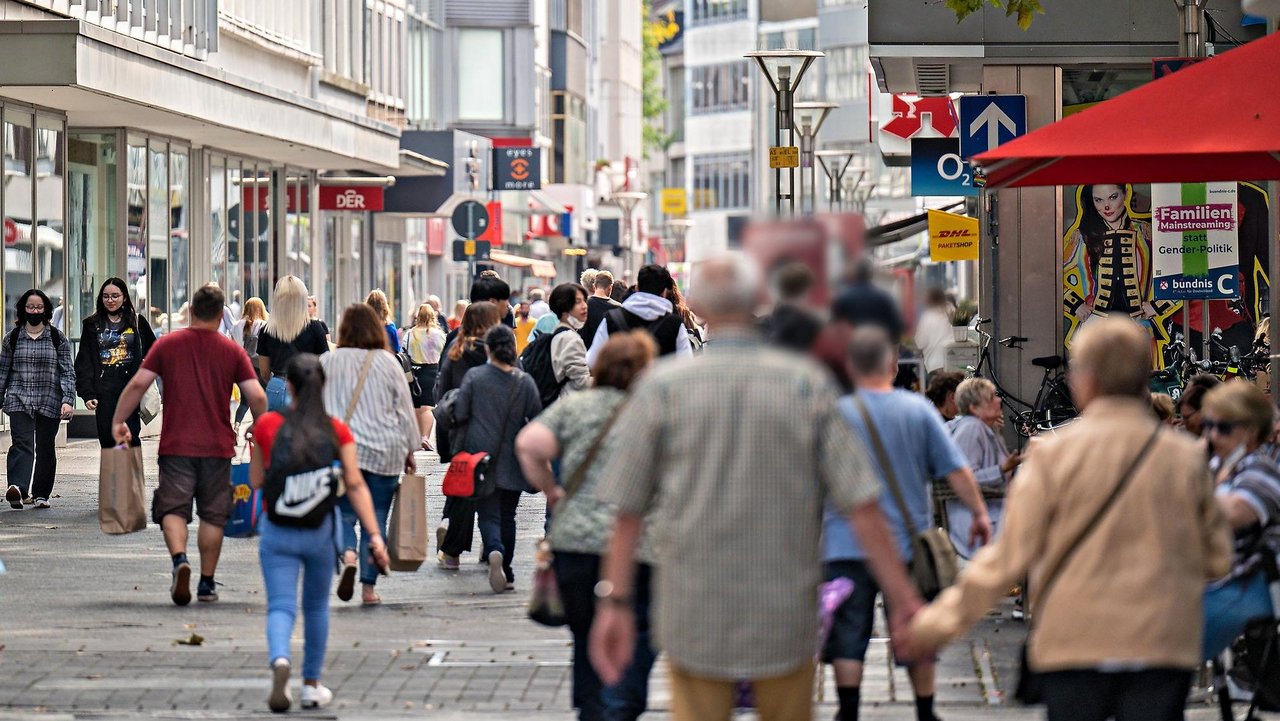 Belebte Einkaufsstraße in der Krefelder Innenstadt mit vielen Passanten. Ältere Menschen im Vordergrund, jüngere im Hintergrund. Läden mit auffälligen Werbeschildern säumen die Straße. Rechts im Bild ein Café mit roten Sonnenschirmen. Die Szene zeigt geschäftiges, urbanes Alltagsleben.