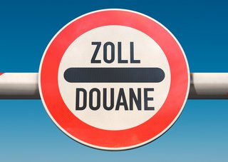 Schranke mit rundem Schild an einem Grenzübergang, Aufschrift „Zoll / Douane“ unter blauem Himmel.