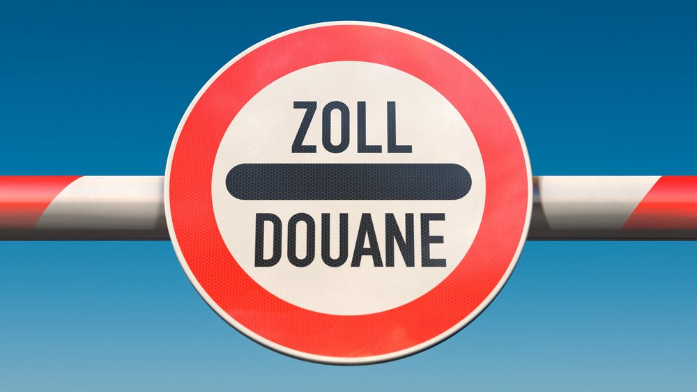 Schranke mit rundem Schild an einem Grenzübergang, Aufschrift „Zoll / Douane“ unter blauem Himmel.