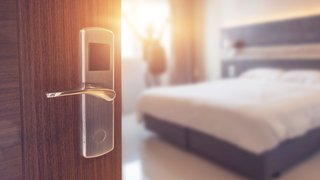 Geöffnete Zimmertür mit elektronischem Türschloss in einem modernen Hotel; im Hintergrund ein helles, aufgeräumtes Schlafzimmer mit Doppelbett und eine Person mit ausgestreckten Armen vor dem Fenster, unscharf dargestellt im Sonnenlicht.