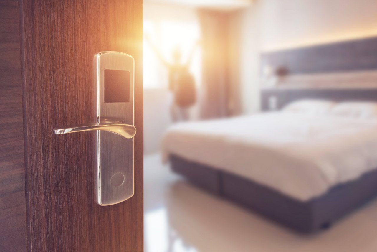 Geöffnete Zimmertür mit elektronischem Türschloss in einem modernen Hotel; im Hintergrund ein helles, aufgeräumtes Schlafzimmer mit Doppelbett und eine Person mit ausgestreckten Armen vor dem Fenster, unscharf dargestellt im Sonnenlicht.