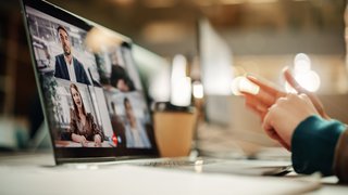 Person zeigt auf einen Laptop mit laufender Videokonferenz, auf dem mehrere Teilnehmende in Kacheln dargestellt sind.
