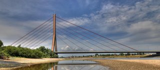 Die Fleher Brücke in Düsseldorf spannt sich bei Tageslicht über den Rhein. Der Wasserstand ist niedrig, wodurch das steinige Flussbett sichtbar wird. Der Himmel ist leicht bewölkt, das Wasser spiegelt die Brücke.