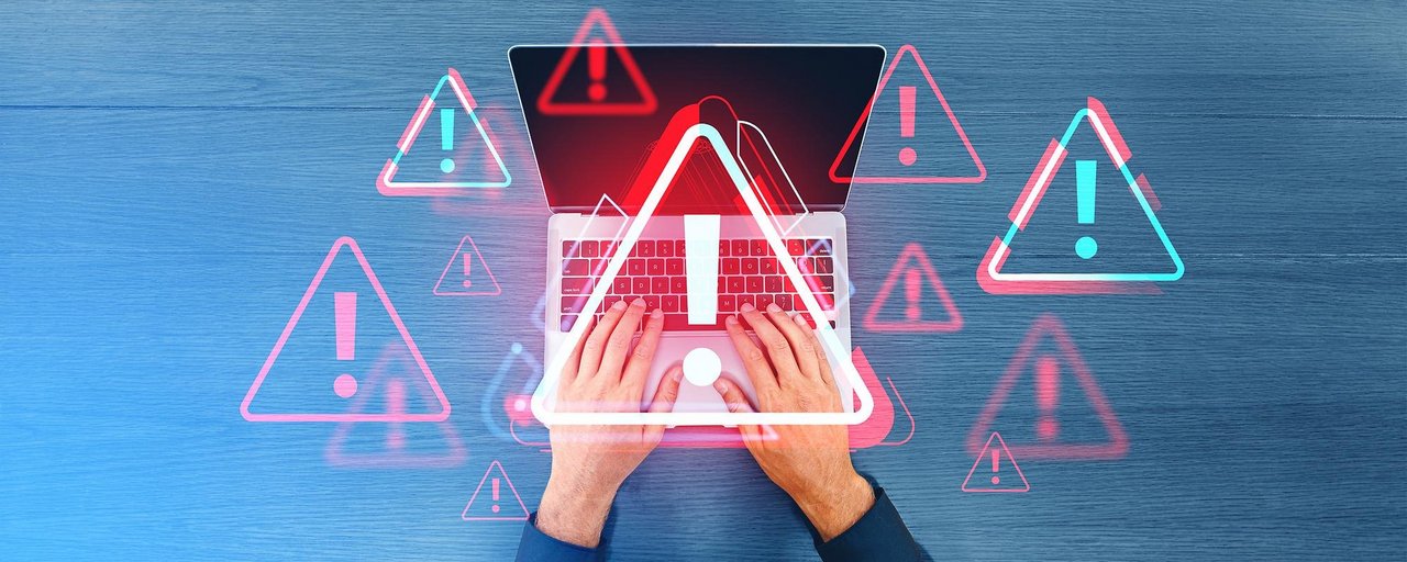 Zwei Hände tippen auf einer Laptop-Tastatur, überlagert von mehreren roten und blauen Warnsymbolen mit Ausrufezeichen; Darstellung symbolisiert eine IT-Sicherheitswarnung oder einen Cyberangriff.
