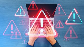 Zwei Hände tippen auf einer Laptop-Tastatur, überlagert von mehreren roten und blauen Warnsymbolen mit Ausrufezeichen; Darstellung symbolisiert eine IT-Sicherheitswarnung oder einen Cyberangriff.